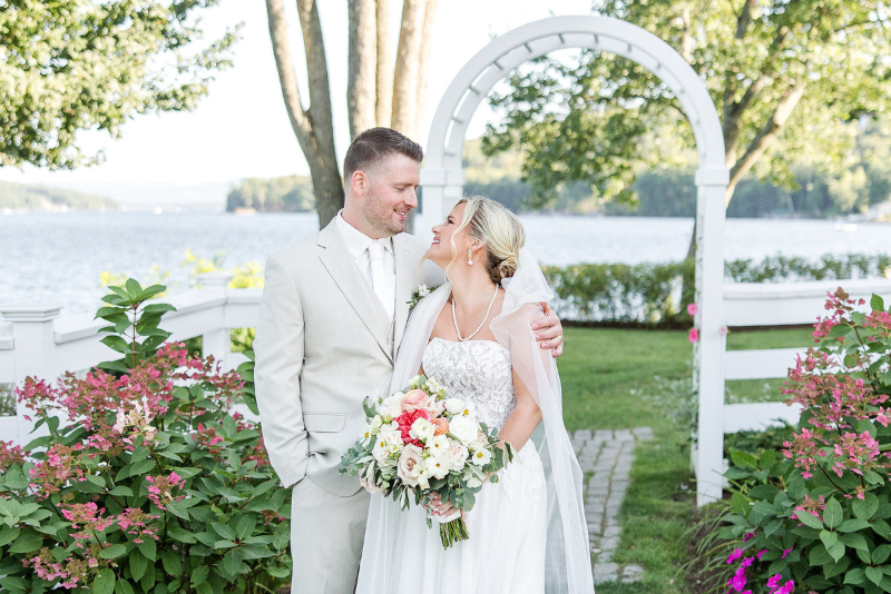 bride and groom pose in front of flowers and garden, with white arch and lake in background